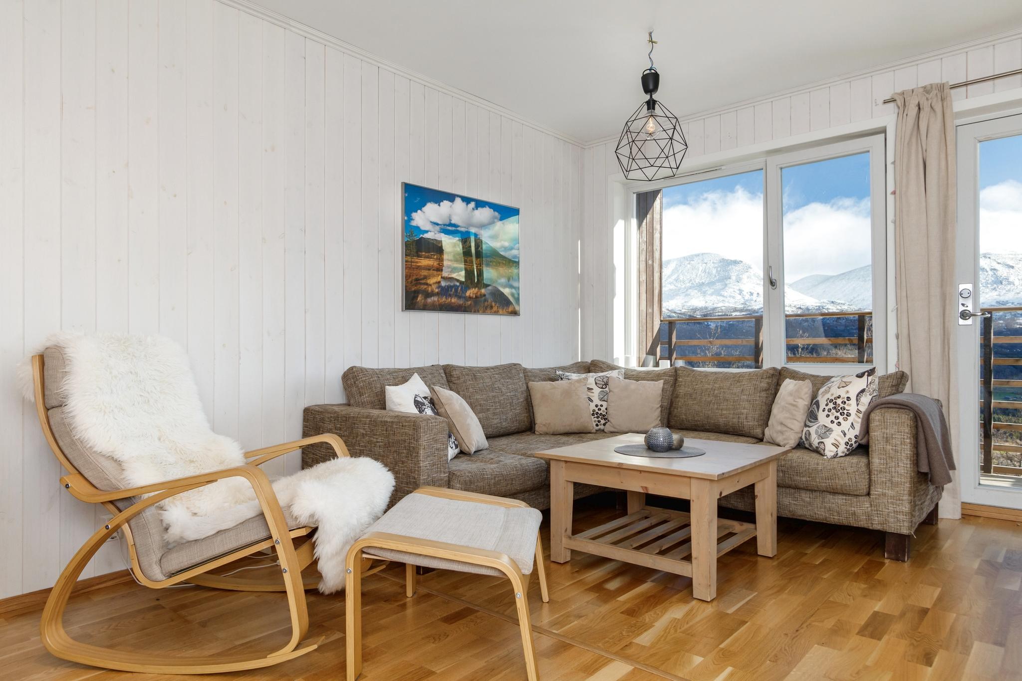 En hyggelig stue, der minder om en charmerende Westendorf-hytte, med en brun sektionssofa med diverse puder, et sofabord i træ og en lys gyngestol i træ med fodstøtte. Et stort vindue afslører en sneklædt udsigt over bjergene. Væggene er hvide, og en geometrisk pendel hænger over hovedet.