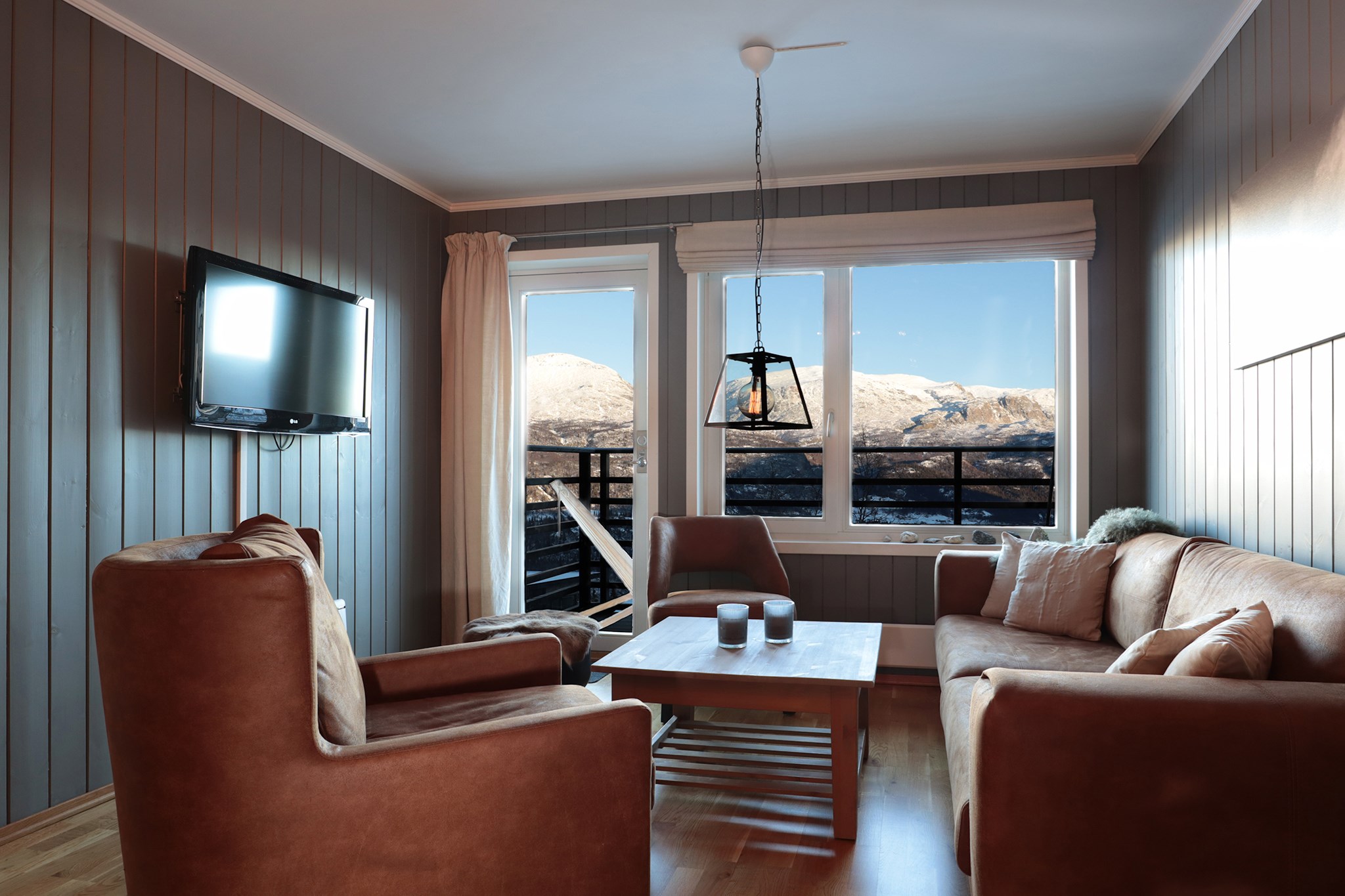 En hyggelig stue med et brunt sofasæt, et sofabord og et væghængt tv. Store vinduer tilbyder en naturskøn udsigt over Wagrains sneklædte bjerge og en balkon med et gelænder. Rummet har trægulve og grå panelvægge, perfekt til afslapning efter skirejseeventyr.