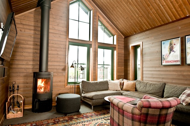 En hyggelig stue med trævægge og -loft, der minder om en hytte i Westendorf, har en hjørnesofa med plaid tæppe, en rund skammel, en tændt brændeovn og et stort vindue. Indrammet kunst og et mønstret tæppe tilføjer varme til rummet, perfekt til skirejseentusiaster.