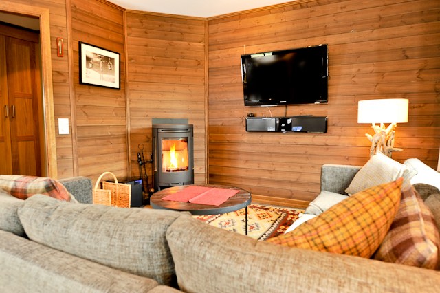 En hyggelig stue med trævægge ligner en hytte, du finder på en skiferie. Den har en moderne pejs, vægmonteret tv, rundt sofabord og grå sektionssofa prydet med plaidpuder. En varm lampe med en unik træbase tilføjer omgivende lys til scenen.