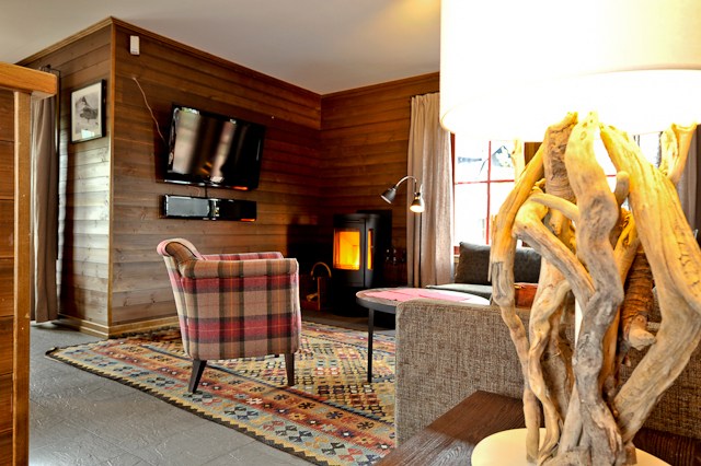 En hyggelig stue med trævægge fremkalder Saalbachs alpine charme, med et mønstret tæppe, en plaid lænestol og en brændeovn. Et fladskærms-tv er monteret på væggen, mens naturligt lys lyser rummet op gennem gardiner, der minder om en Wagrain sommerferie.
