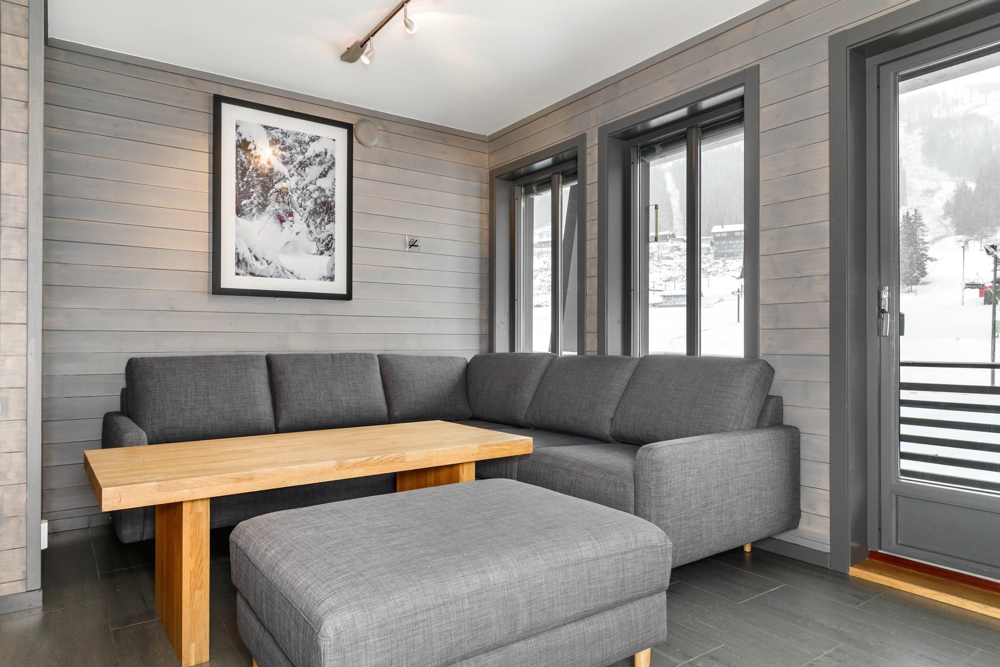 En moderne stue med grå vægge og en stor grå sektionssofa fremkalder den fredfyldte charme ved en Wagrain skiferie. Et sofabord i træ og matchende skammel sidder foran sofaen, mens et indrammet naturfotografi pryder væggen. Store vinduer indrammer et snelandskab, der minder om Saalbachs vintertiltrækning.