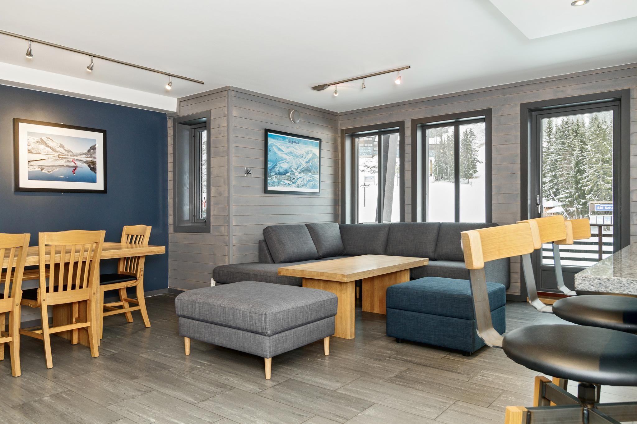 Et moderne opholds- og spiseområde har et spisebord i træ med stole, en grå sektionssofa og en skammel. Rummet, der er perfekt til et skiferie-retreat, er prydet med malerier på træbeklædte vægge. Store vinduer afslører sneklædte træer udenfor mod gråt klinkegulv.