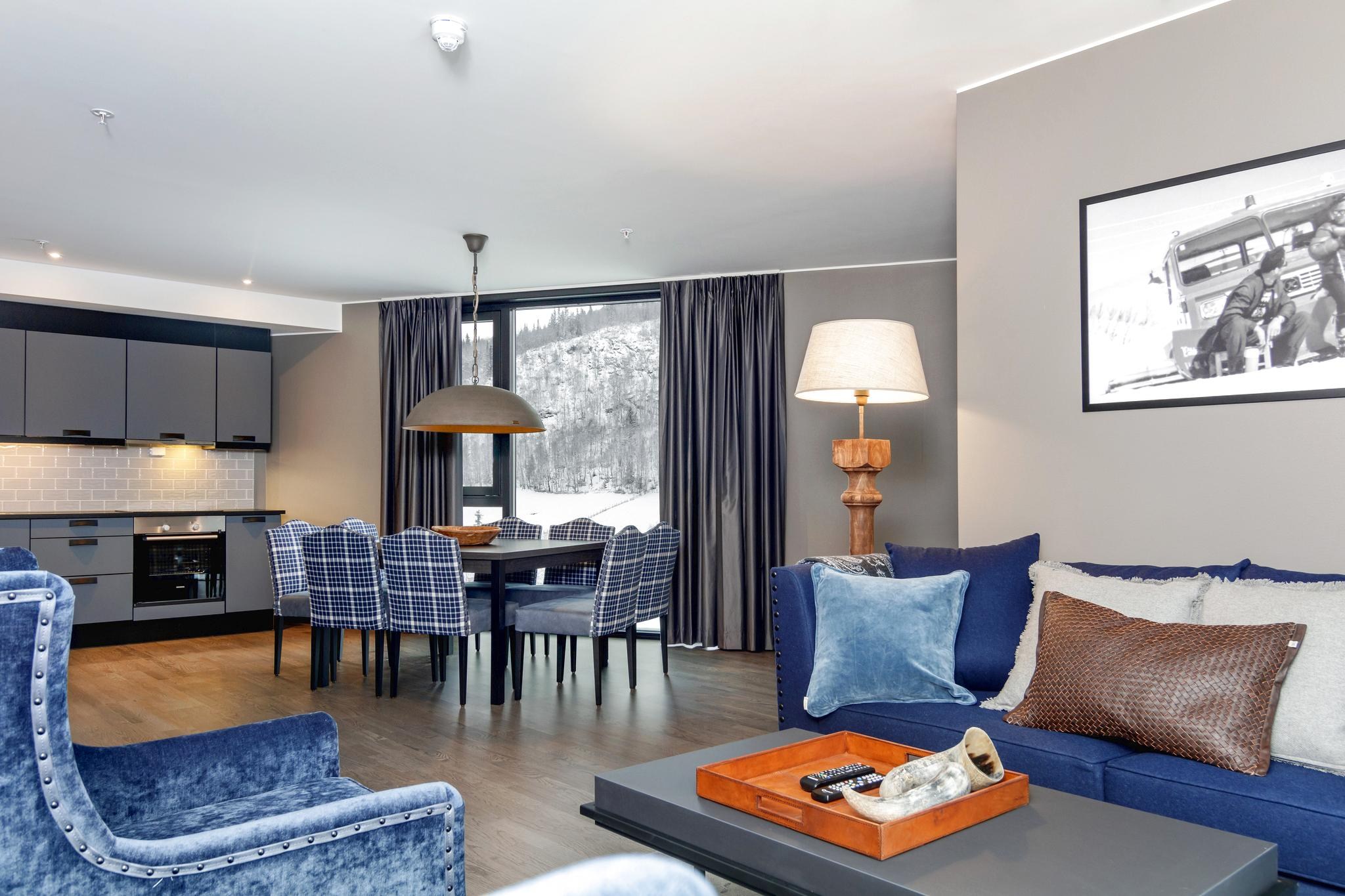 Moderne stue med blå fløjlsstole og sofa, dekorative puder og et sofabord i træ. Et spisebord med blå plaid stole er i nærheden af et vindue, der viser en fantastisk udsigt over bjergene, der minder om Saalbach. En gulvlampe og et indrammet foto tilføjer stemning. Køkkenet har mørke skabe.