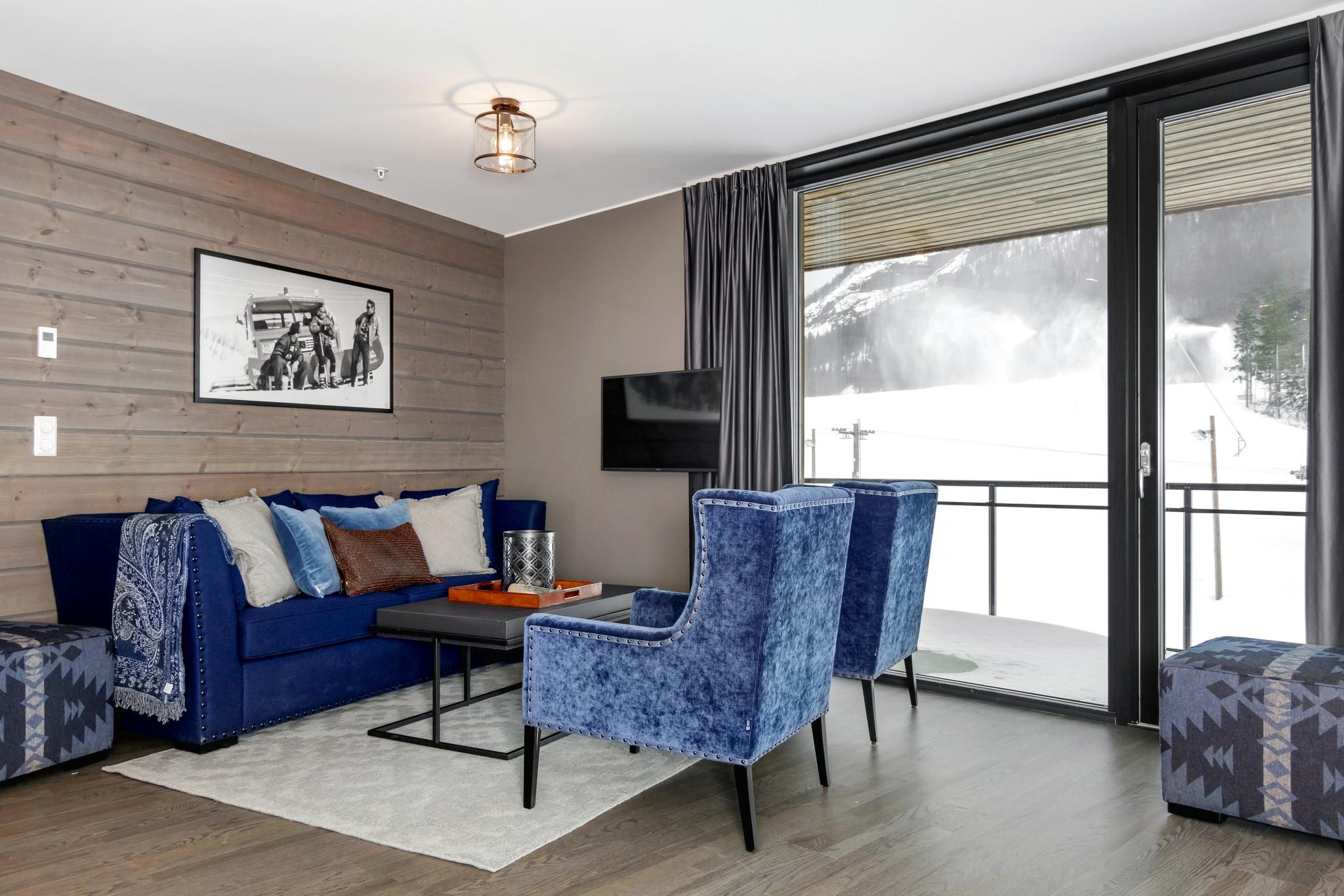 En moderne stue i Westendorf har en blå sofa, to blå lænestole og et lille sofabord. Et vægmonteret tv sidder overfor store vinduer, der afslører sneklædte bjergudsigter. Indretningen omfatter et mønstret tæppe og et indrammet sort-hvidt foto på væggen.