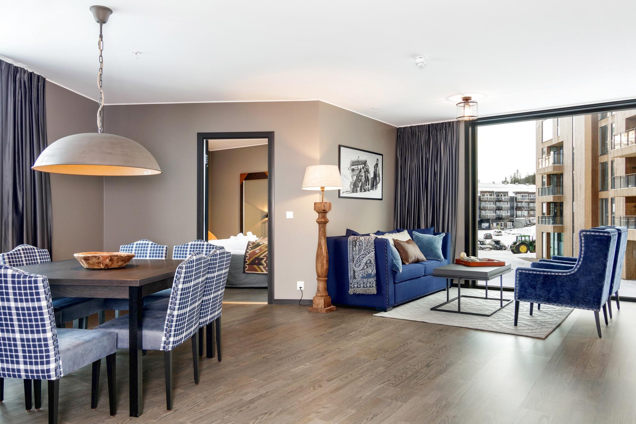 Et moderne opholds- og spiseområde med trægulve, der gentager Saalbachs charme. Værelset har et spisebord med seks ternede stole, en blå sofa prydet med hynder, en stor gulvlampe, kunst på væggene og store vinduer, der giver naturligt lys og udsigt til bygningerne udenfor.