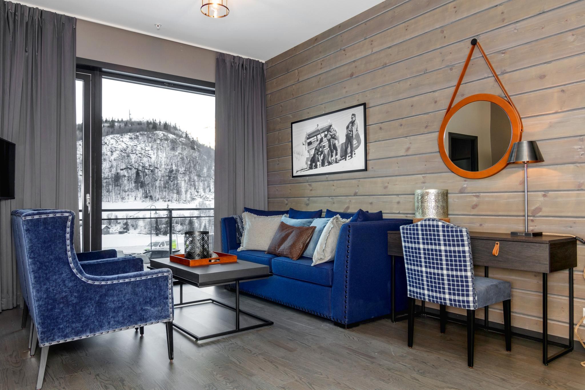 En hyggelig stue i hjertet af Saalbach har en blå sofa med farverige hynder, to blå lænestole, et sofabord i træ og et skrivebord med en lampe. Et rundt orange spejl pryder panelvæggen, mens vinduet indrammer en betagende udsigt over sneklædte bjerge.