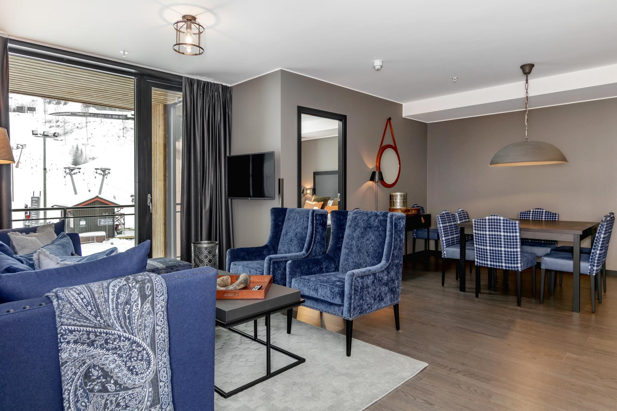 Moderne stue med blå fløjlsstole og en sofa, et fladskærms-tv på væggen og et spisebord med plaidstole. Rummet, der er perfekt til at mindes om Wagrain skirejser, har store vinduer med udsigt over bjergene og mørke gardiner, suppleret med stilfulde lysarmaturer.