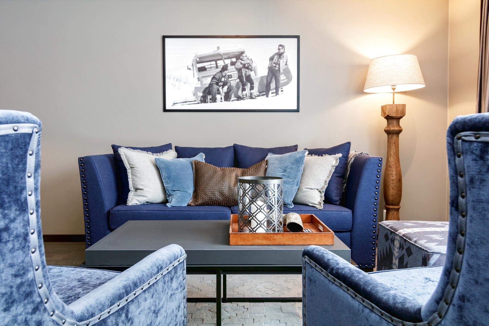 En hyggelig stue med en blå sofa prydet med beige og blå puder, der minder om en afslappende sommerferie i Østrig. Et indrammet sort-hvidt foto hænger over sofaen. To blå lænestole og en trælampe flankerer et elegant sofabord med en bakke og pynteting.