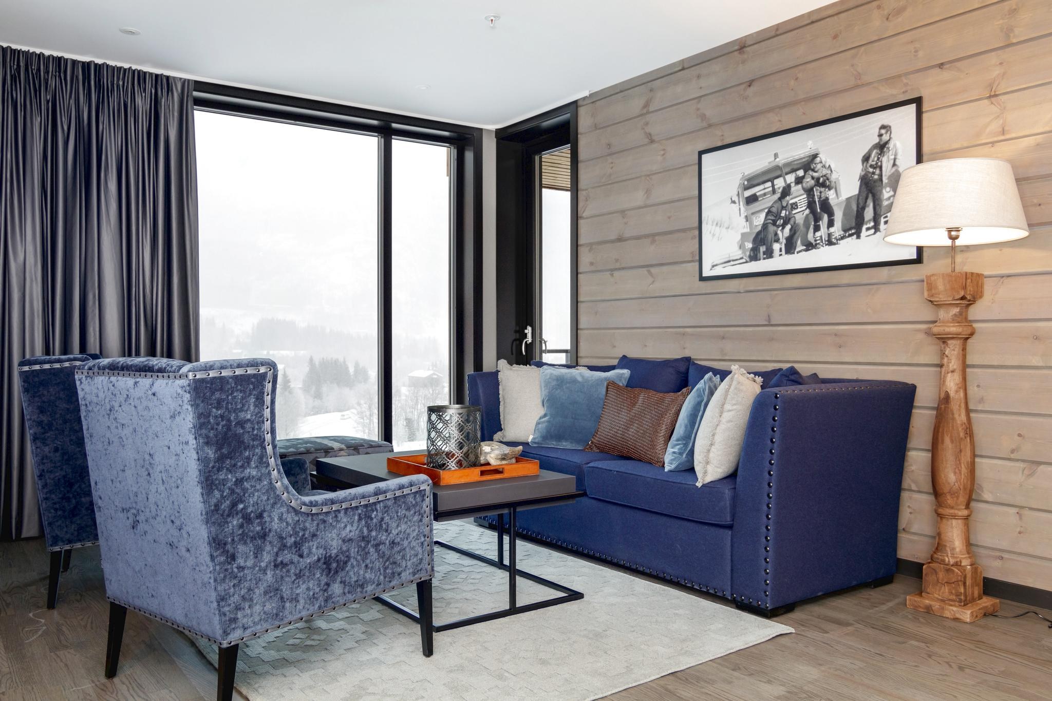En moderne stue med en blå sofa, to matchende lænestole og et mørkt sofabord fremhæves af et vintage sort-hvidt foto. Store vinduer med mørke gardiner afslører Saalbachs snelandskab udenfor, hvilket skaber den perfekte flugt for skirejseentusiaster.