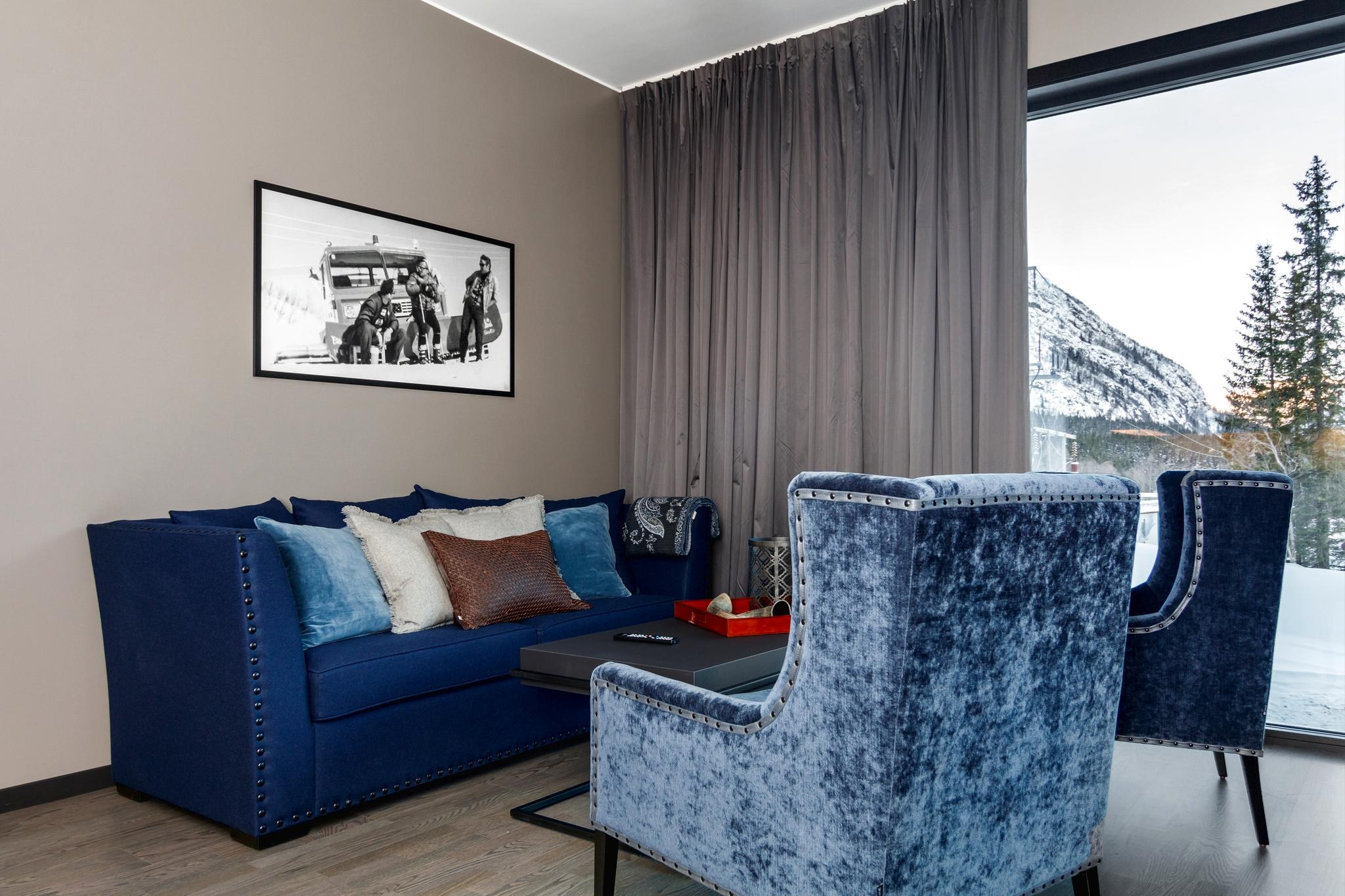 En moderne stue har en blå sofa og to fløjlslænestole omkring et sort sofabord. Et monokromt fotografi hænger på væggen med store vinduer, der afslører sneklædte bjerge og høje træer - perfekt til at mindes om din skiferie i Saalbach.
