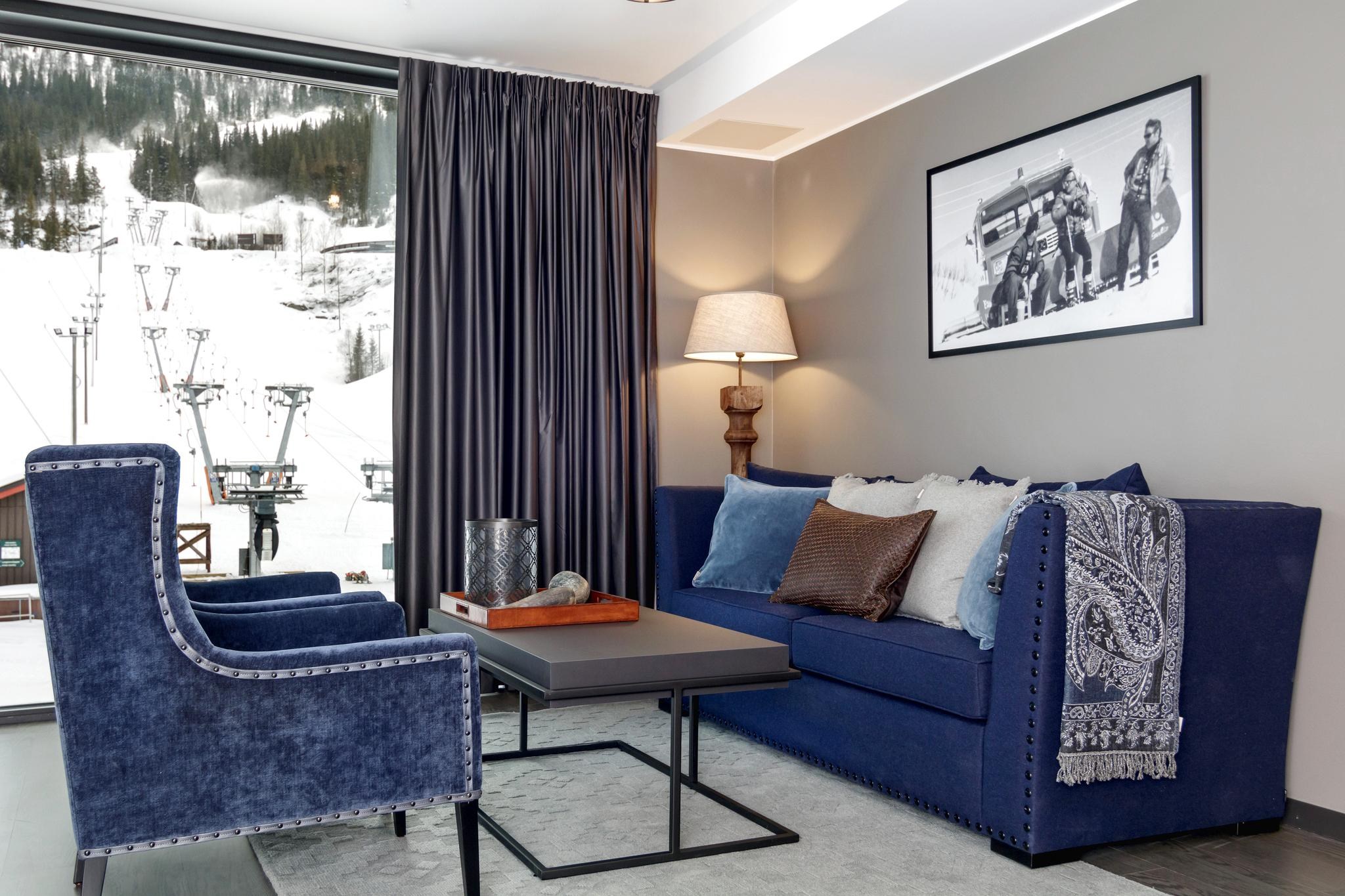 En stilfuld stue i Westendorf-inspirerede omgivelser har en blå sofa og lænestole, et mønstret træk og et indrammet sort-hvidt foto på væggen. En gulvlampe og et sofabord med dekorationer fuldender det hyggelige setup, mens vinduet afslører en sneklædt skiliftscene udenfor.