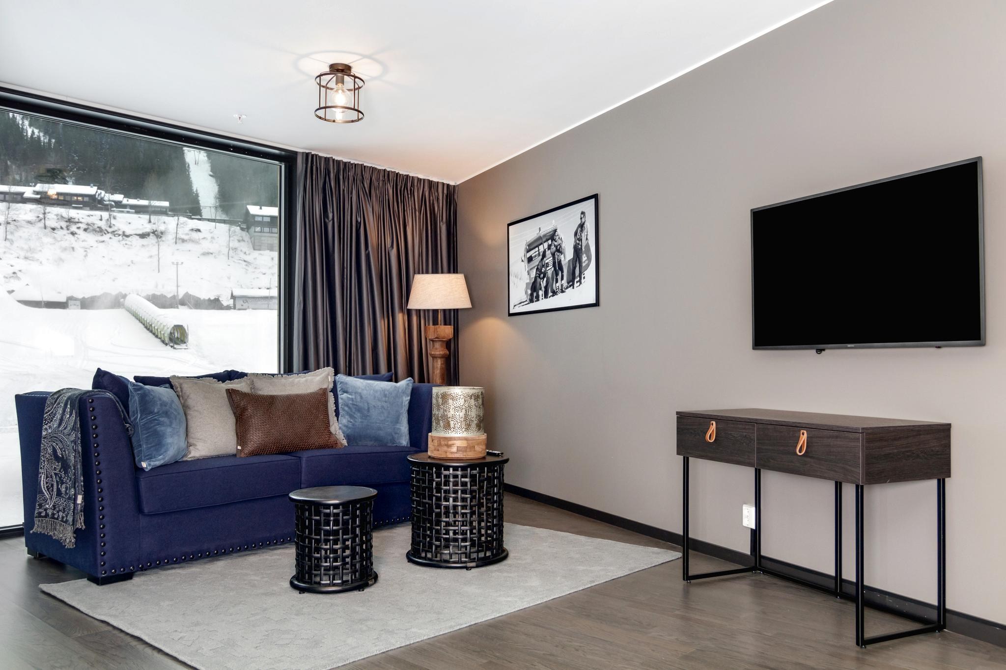 Den moderne stue, der minder om et hyggeligt Saalbach-retræte, har en marineblå sofa med brune puder og to sorte sideborde på et lyst tæppe. Et fladskærms-tv er monteret over et elegant konsolbord. Store vinduer viser en sneklædt udendørs udsigt, mens loft- og gulvlamper byder på rigelig belysning.