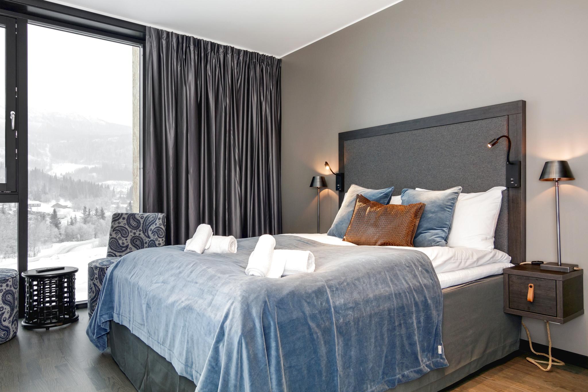 Et moderne hotelværelse i Westendorf har en stor seng med blåt sengetøj og puder. Rullede håndklæder sidder på sengen, mens store vinduer med mørke gardiner fremviser et vinterlandskab perfekt til en skiferie. To natborde med lamper flankerer sengen, klar til at hilse på dit hyggelige tilflugtssted.