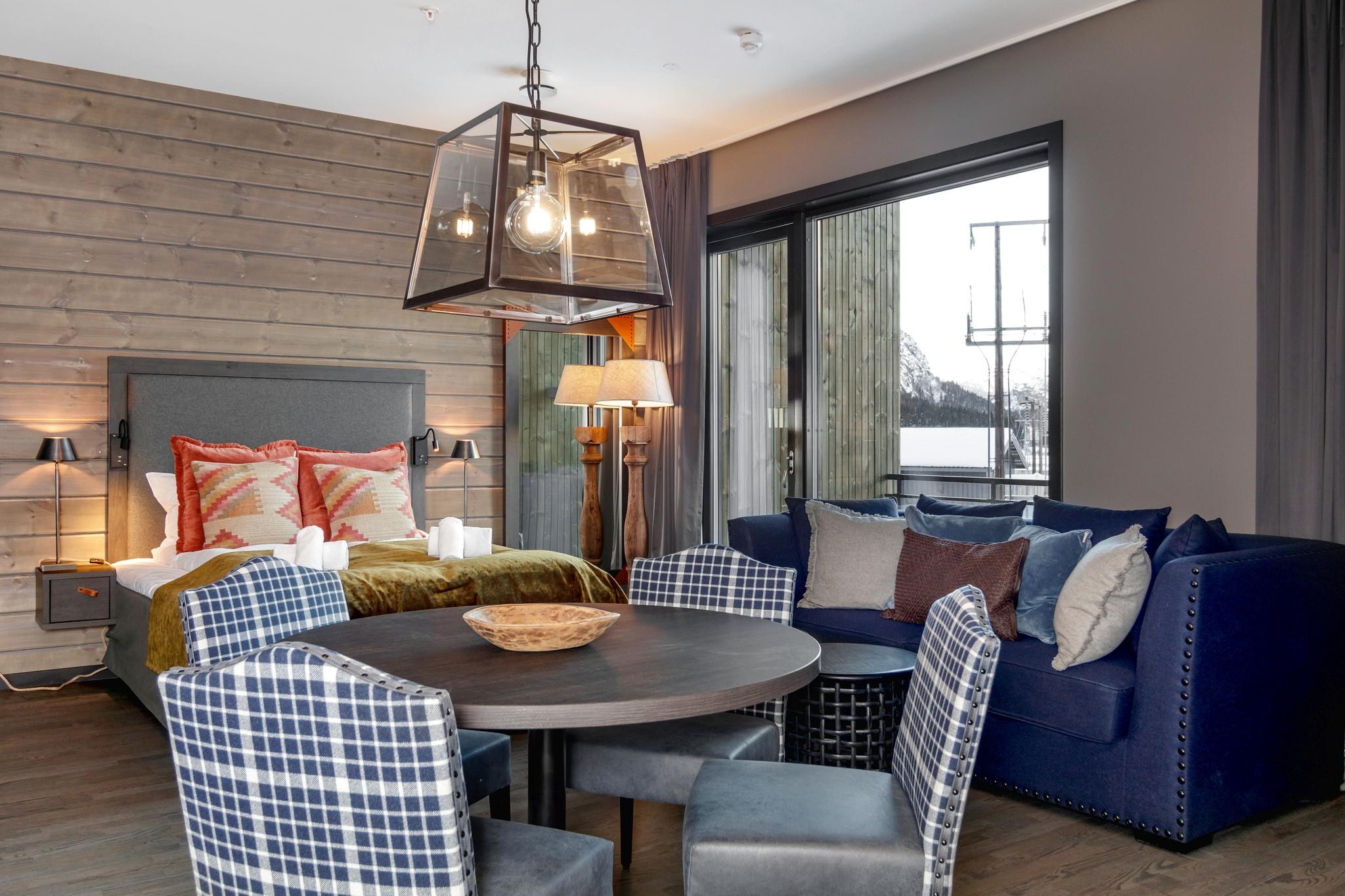 Et hyggeligt hotelværelse i Wagrain har en stor seng med farverige puder og et blødt puds. I nærheden supplerer et rundt bord med fire plaid-stole den marineblå sofas elegance. Store vinduer giver udsigt over snelandskabet, perfekt for skirejse-entusiaster. Varm belysning skaber en indbydende stemning.