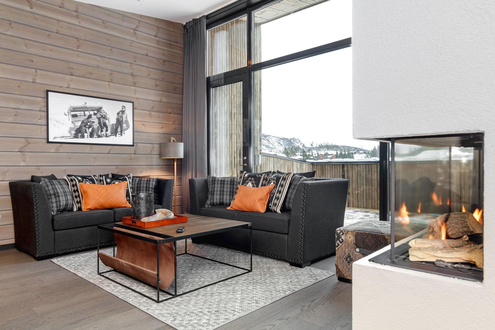 En hyggelig stue i Wagrain har mørke sofaer med orange og plaidpuder. Et sofabord i træ står på et mønstret tæppe, mens et indrammet foto hænger på vægge med træpaneler. En moderne pejs og store vinduer afslører det naturskønne snelandskab på denne perfekte skiferiedestination.