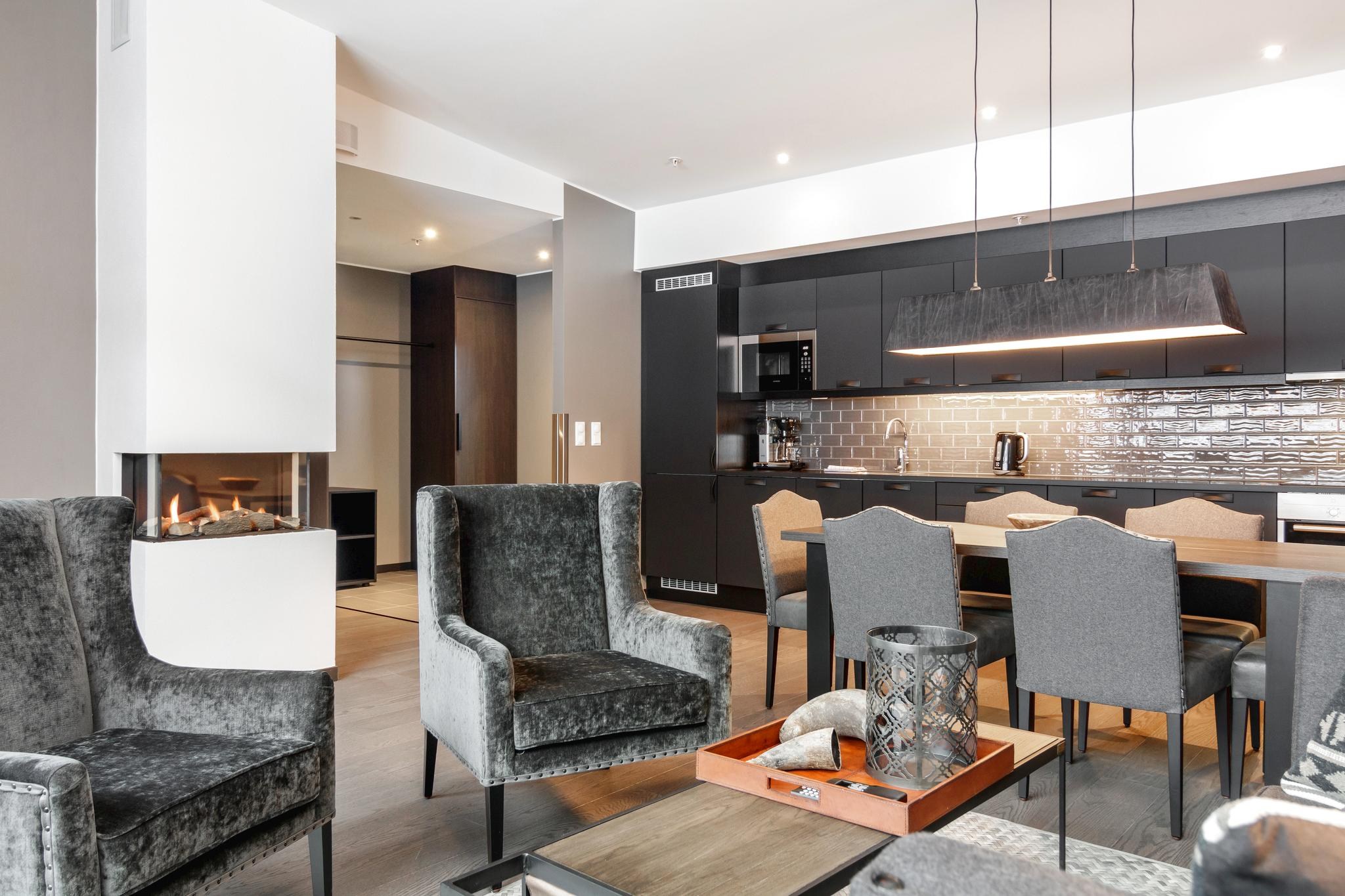 Et moderne opholds- og spiseområde i åbent plan med en hyggelig atmosfære, der minder om en Wagrain skirejser. Den har en pejs, fløjlslænestole, et spisebord med grå stole og et elegant køkken med mørke skabe og en murstensplade. En dekorativ bakke står på sofabordet.