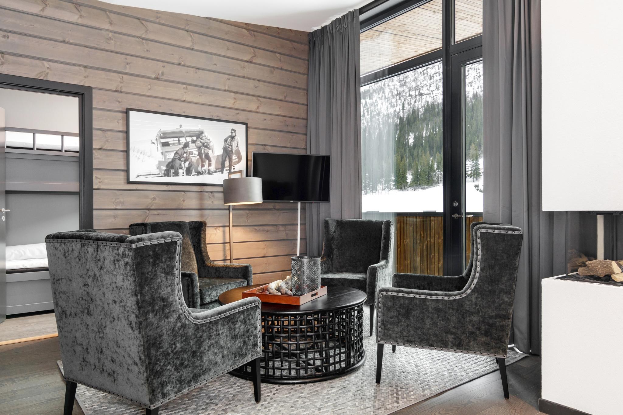 Moderne stue med fire grå fløjlslænestole arrangeret omkring et rundt sofabord. Store vinduer afslører den sneklædte bjergudsigt over Saalbach. Et sort-hvidt foto hænger på væggen med træpaneler, og mørke gardiner indrammer vinduerne.