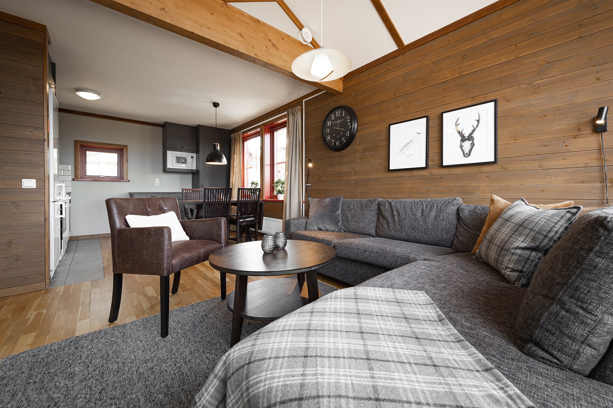 En hyggelig stue med trævægge og højt til loftet, der minder om en Saalbach skiferie, byder på en grå sektionssofa med plaidpuder, et rundt sofabord i træ og en mørk lænestol. Kunst og et ur pryder væggene. En spiseplads og køkken er synlige i baggrunden.