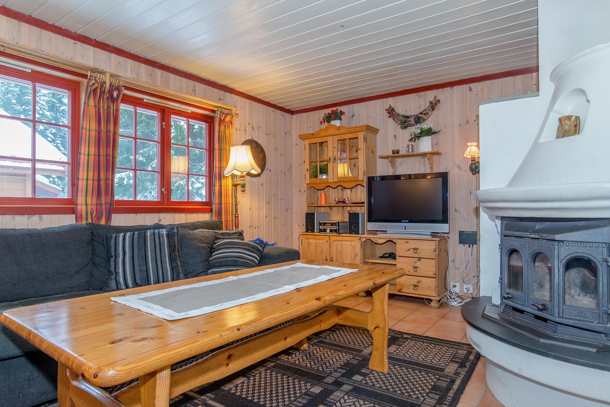 En hyggelig stue med træbeklædte vægge fremkalder charmen ved en Wagrain skiferie. Den har en grå hjørnesofa, træsofabord og et tv på et træskab. En hvid pejs står til højre, mens plaidgardiner indrammer vinduet og indbyder til naturligt lys.