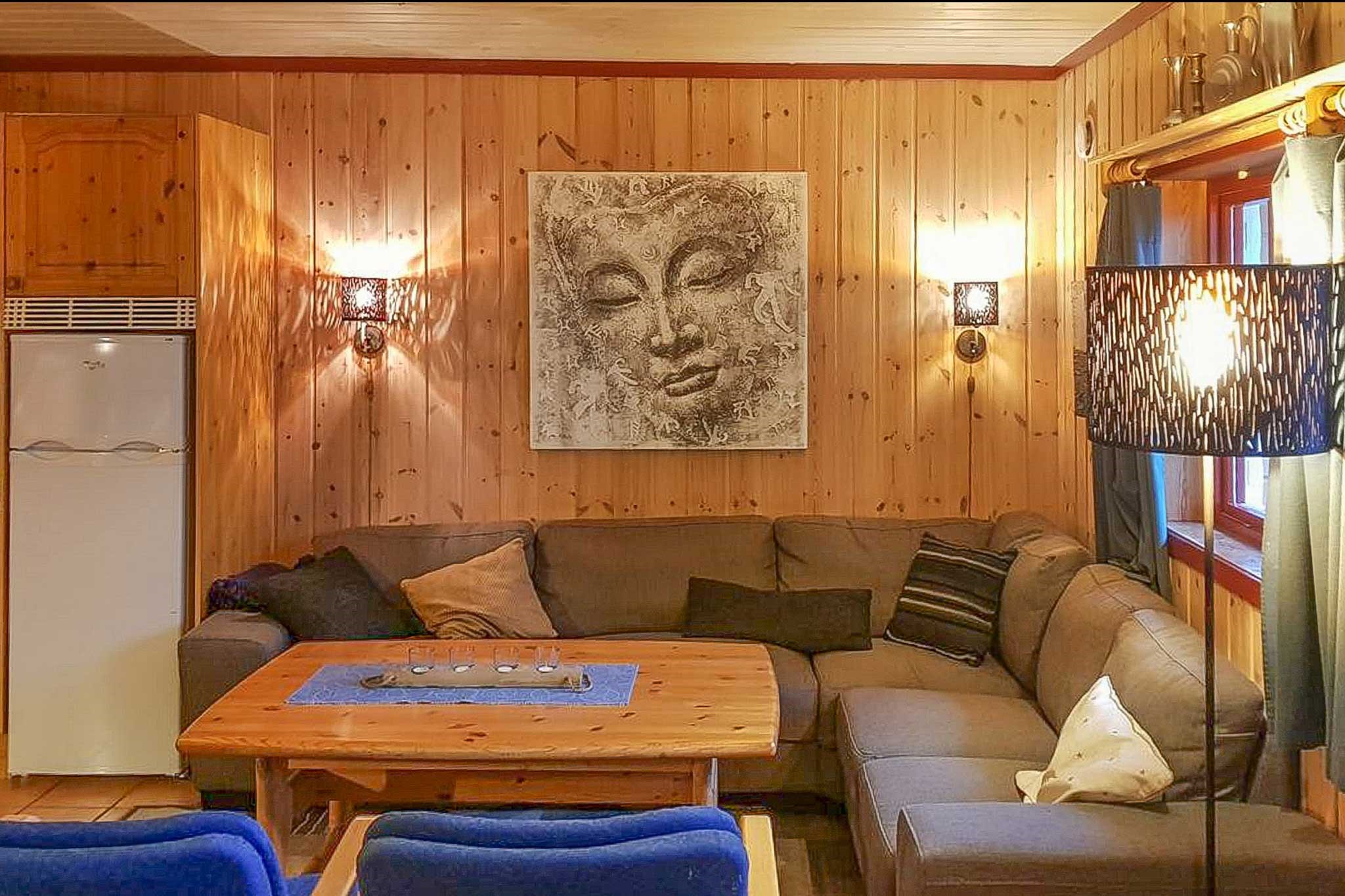 En hyggelig stue med træpaneler, der minder om en skiferie i Wagrain, har en stor L-formet sofa med diverse hynder. Et sofabord i træ er i centrum, og et fredfyldt ansigtskunstværk hænger over sofaen. Blød belysning fuldender den varme, indbydende atmosfære.