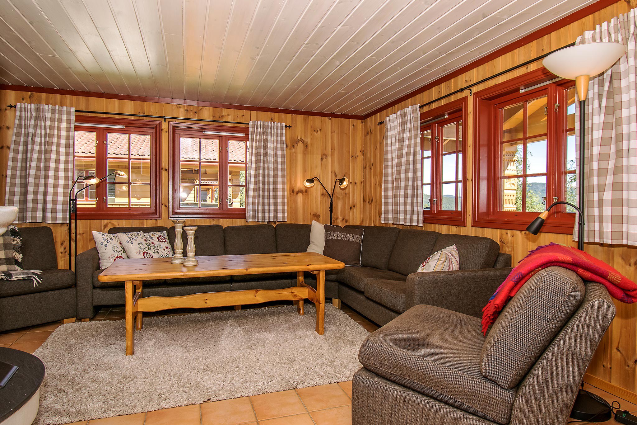 En hyggelig stue med trævægge og loft, der minder om en Wagrain-hytte. Den har en stor, brun sektionssofa med diverse hynder, et sofabord i træ og et blødt beige tæppe. Vinduer med plaidgardiner lukker naturligt lys ind, og et par gulvlamper er til stede.
