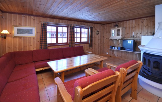 En hyggelig stue med trævægge og loft, der minder om et Westendorf-retræte. Den har en stor rød L-formet sofa, to matchende stole og et sofabord i træ. En hvid pejs og et TV med stativ er i hjørnet. Ternede gardiner pryder vinduerne og indbyder til tanker om skirejseaftener.
