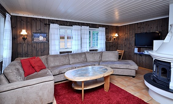 Hyggelig stue med en stor L-formet beige sofa, et rundt sofabord i træ og et rødt tæppe. Inspireret af Saalbachs alpine charme har værelset mørkt træpanel, hvidt loft og gardiner. Et fladskærms-tv er monteret på væggen og et komfur varmer hjørnet.