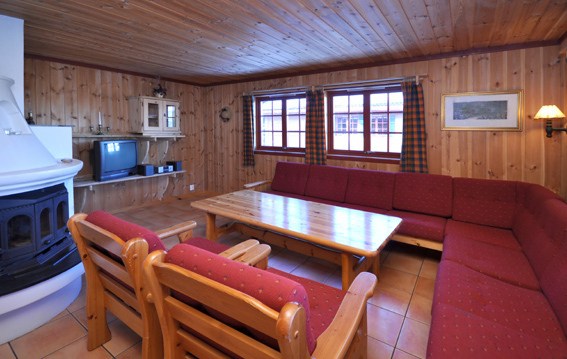 En hyggelig stue med træpaneler på vægge og loft, perfekt til afslapning efter en skiferie i Westendorf. Den har en stor rød sektionssofa, træstole, et sofabord, et fjernsyn på en hylde og en hvid pejs i hjørnet. To vinduer med plaidgardiner lukker naturligt lys ind.
