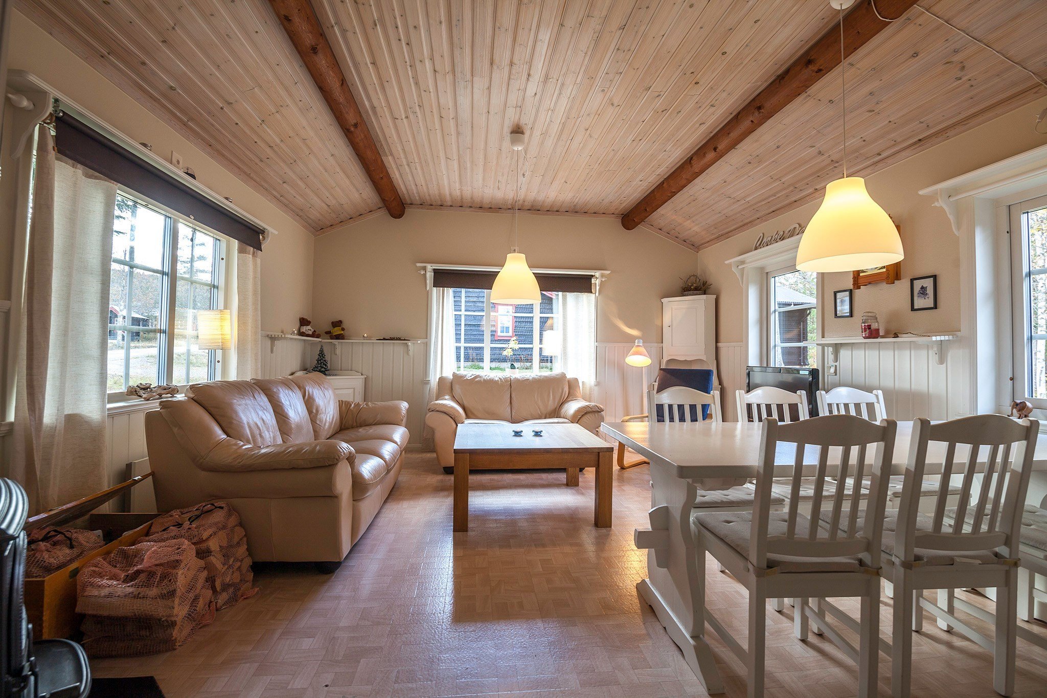 En hyggelig stue, der minder om en Westendorf skihytte, med træloft og parketgulv. Den har to beige sofaer, et sofabord i træ og en spiseplads med et hvidt bord og stole. Store vinduer indleder naturligt lys, mens pendullamper tilføjer varme fra oven.