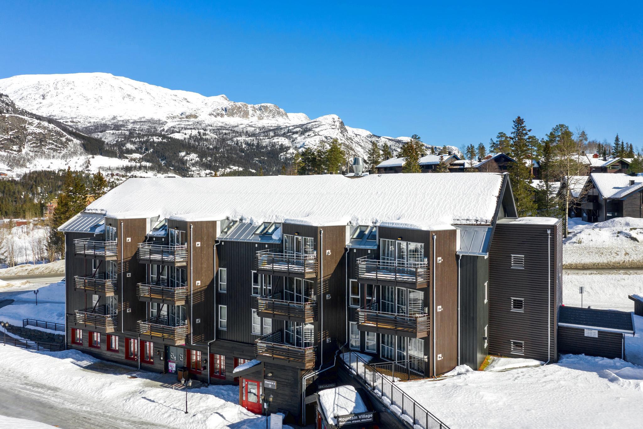 En moderne tre-etagers bygning med balkoner, dækket af sne, står på baggrund af snedækkede bjerge og klar blå himmel - et perfekt tilflugtssted for en skiferie. Det omkringliggende landskab er oversået med træer og andre strukturer, der fanger essensen af et vintervidunderland.