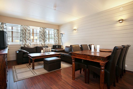 En hyggelig opholds- og spisestue med trægulve og vægge, der minder om et skiferie-retræte. Værelset har en sofa, lænestol, sofabord og et spisebord til otte. Lyset filtrerer gennem mønstrede gardiner, der dækker store vinduer, og forstærker den varme atmosfære, der ligner Westendorfs charme.