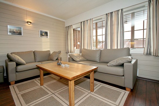 En hyggelig stue, der minder om et Wagrain-hytte med to beige sofaer, et sofabord i træ og mønstrede gardiner. To små indrammede billeder hænger på den lyse panelvæg. Et tæppe med et geometrisk mønster dækker det mørke trægulv, mens naturligt lys strømmer gennem vinduerne.