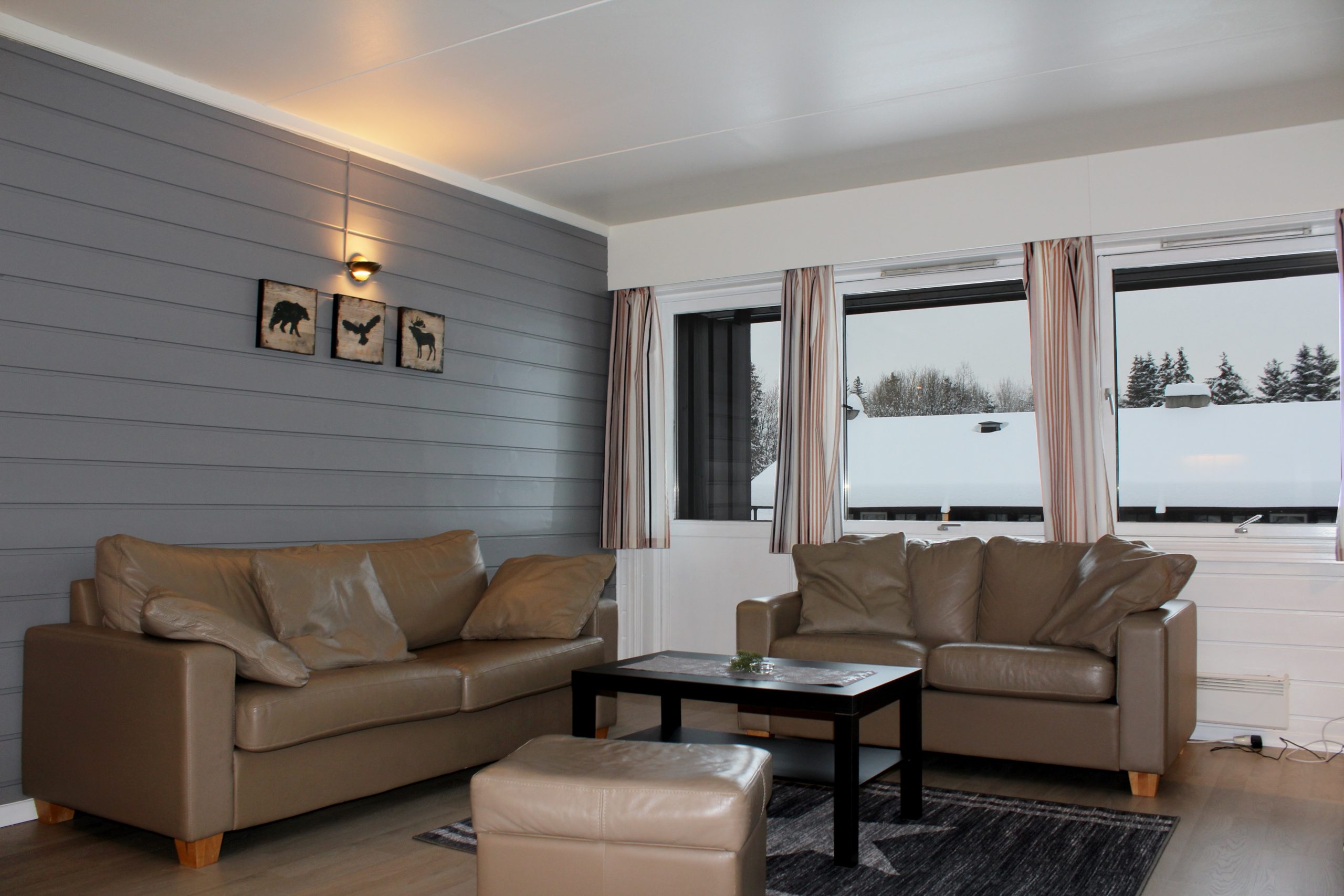 En hyggelig stue med grå panelvægge, et brunt lædersofasæt og et lille sofabord i træ. Tre indrammede dyresilhuetter pryder væggen. Store vinduer afslører et snelandskab, der minder om Wagrain, og beige gardiner indrammer udsigten.