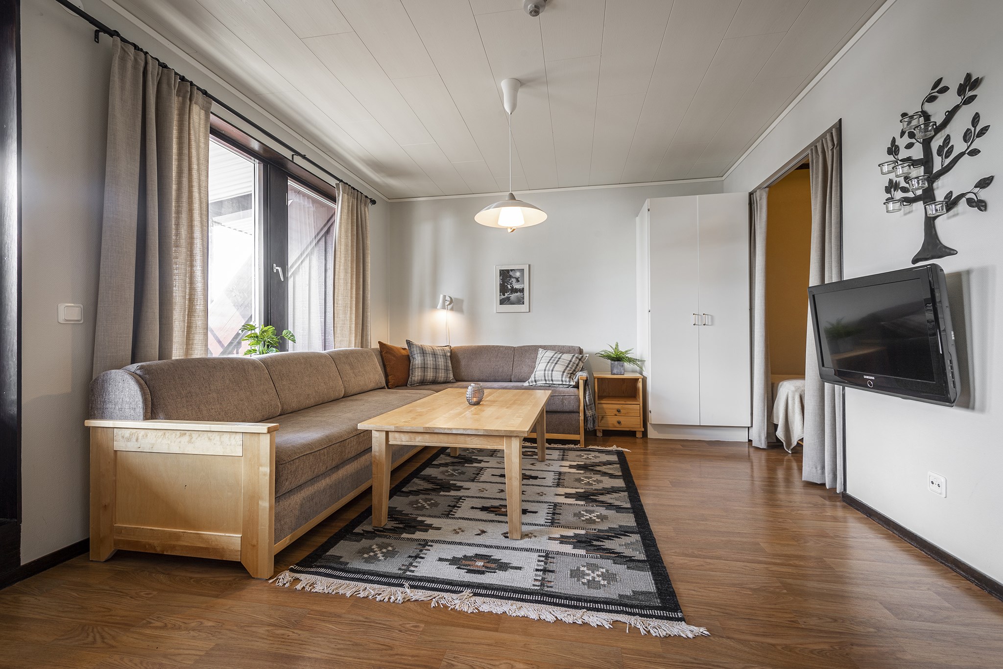 En hyggelig stue, der minder om en Saalbach skiferie, med brun sektionssofa, mønstret tæppe og sofabord i træ. Naturligt lys strømmer gennem store vinduer med beige gardiner. Et maleri hænger over et lille sidebord, og fuldender denne indbydende alpine tilbagetog stemning.