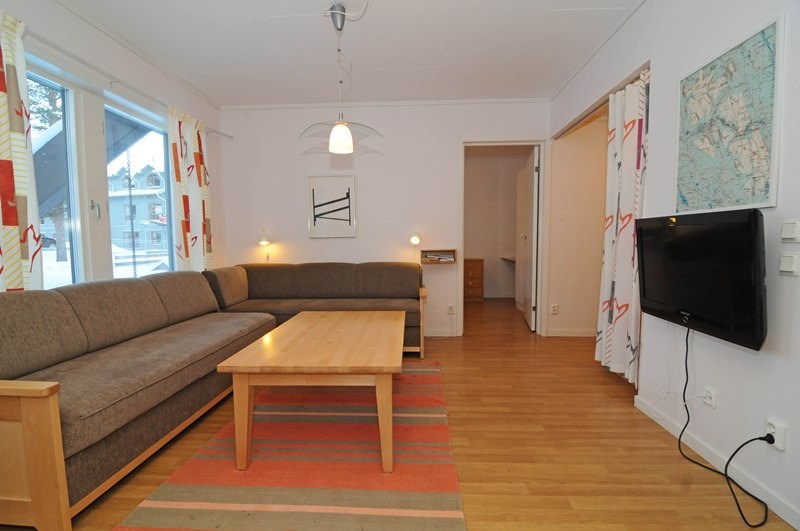 En hyggelig stue med en stor brun sektionssofa, et sofabord i træ og et fladskærms-tv monteret på væggen. Kortvægkunst tilføjer et strejf af vandrelyst, der minder om Westendorf i sommerferie Østrig. Naturligt lys filtrerer gennem mønstrede gardiner ud på trægulvet.
