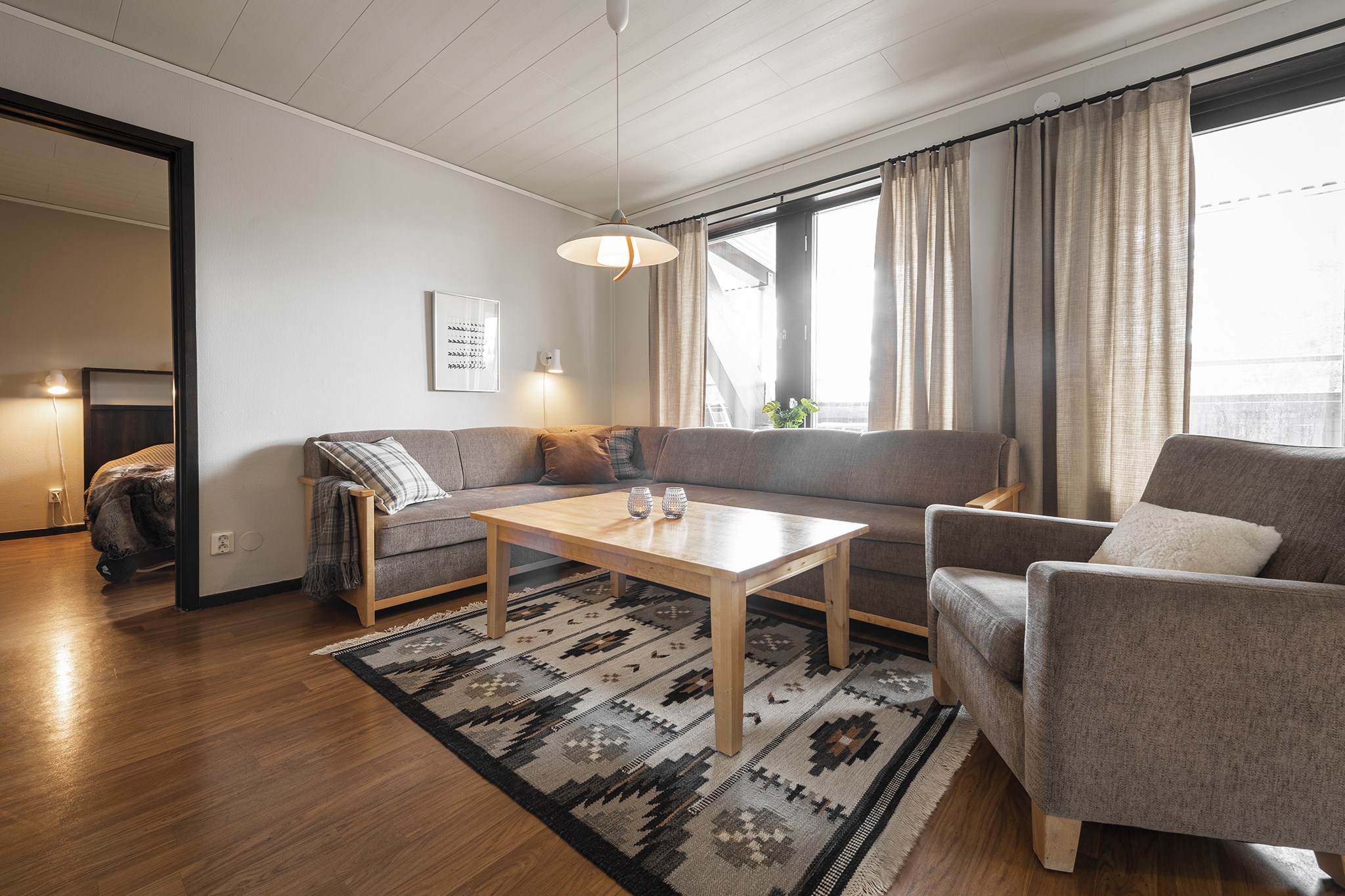 En hyggelig stue med et sofabord i træ, to grå sofaer og en lænestol. Et geometrisk mønstret tæppe pryder trægulvet, mens store vinduer med beige gardiner lukker naturligt lys ind, der minder om et Westendorf-skiferiested. Et soveværelse er synligt gennem en åben dør.