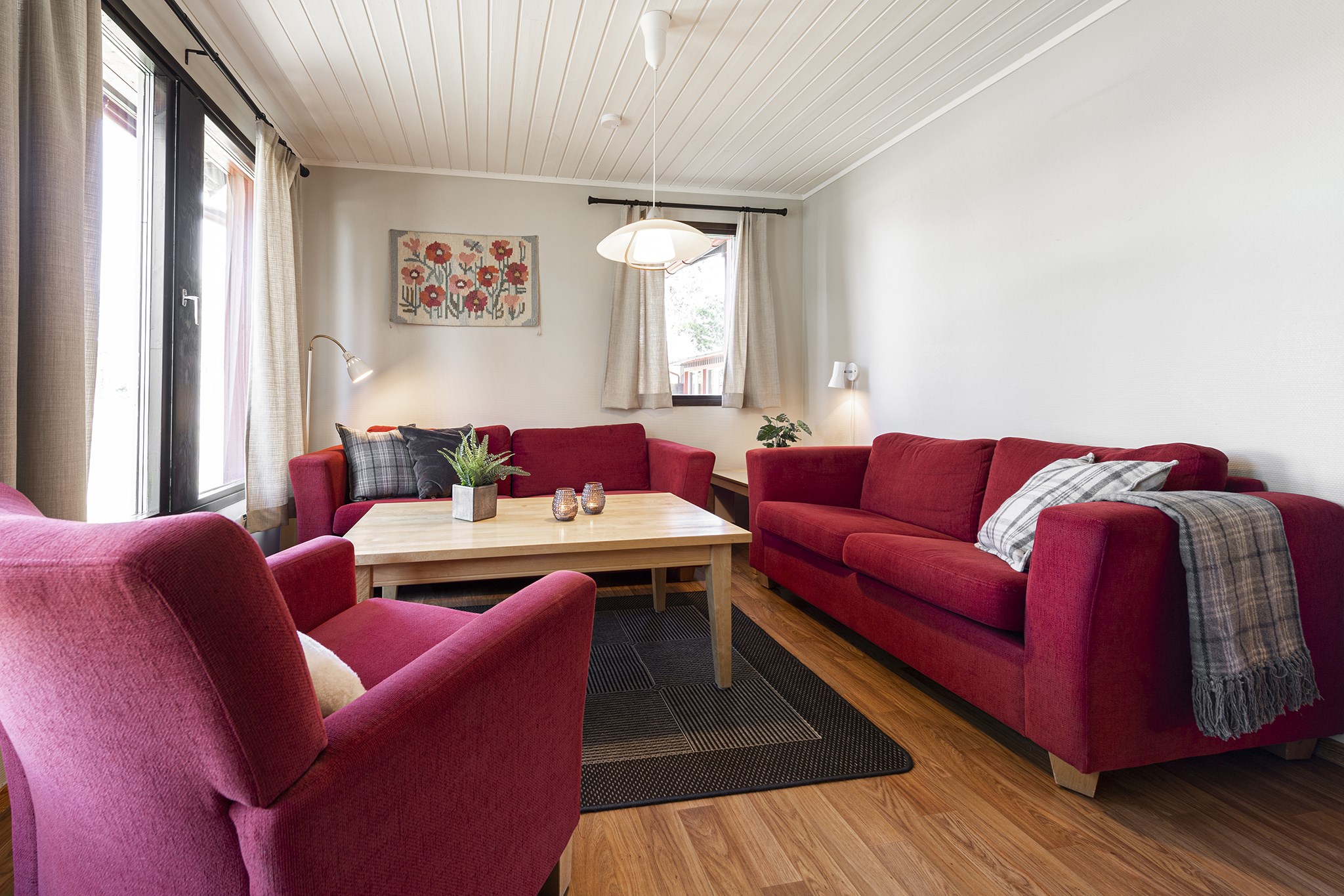 En hyggelig stue i Wagrain med røde sofaer og lænestole arrangeret omkring et sofabord af træ. En gulvlampe står ved siden af siddepladserne, mens et farverigt blomstermaleri pryder væggen. Værelset, perfekt efter skiferieeventyr, har trægulv og loftslampe.