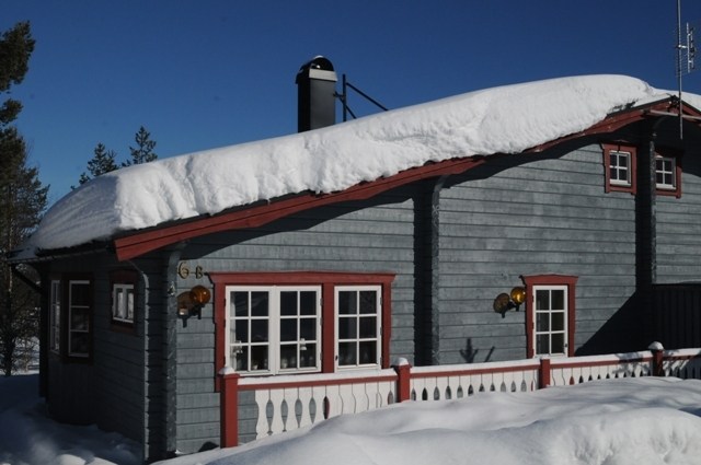 En hytte med blå trævægge og røde trim er dækket af sne, beliggende midt i Wagrains naturskønne skønhed. Tag og altan kan prale af tykke lag af sne under en klar, blå himmel, mens et par træer står elegant bag huset.