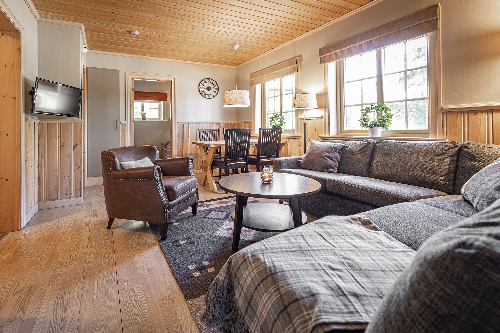 Hyggelig stue med trævægge og loft, der minder om en Wagrain hytte. Med en grå sofa, læderlænestol og rundt sofabord er den perfekt til afterski-afslapning. Et TV er monteret på væggen, og et spisebord med stole er i baggrunden. Store vinduespartier bringer naturligt lys ind.