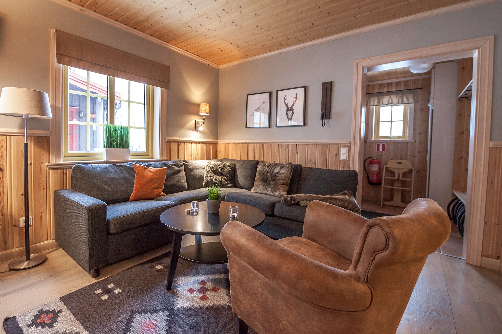 En hyggelig stue, der minder om et charmerende Westendorf-retræte, med en stor grå sektionssofa, en brun lænestol og et rundt sort sofabord. Trævægge og -loft tilføjer til varmen, suppleret med indrammet kunst og et mønstret tæppe. Blød belysning forstærker den indbydende atmosfære.