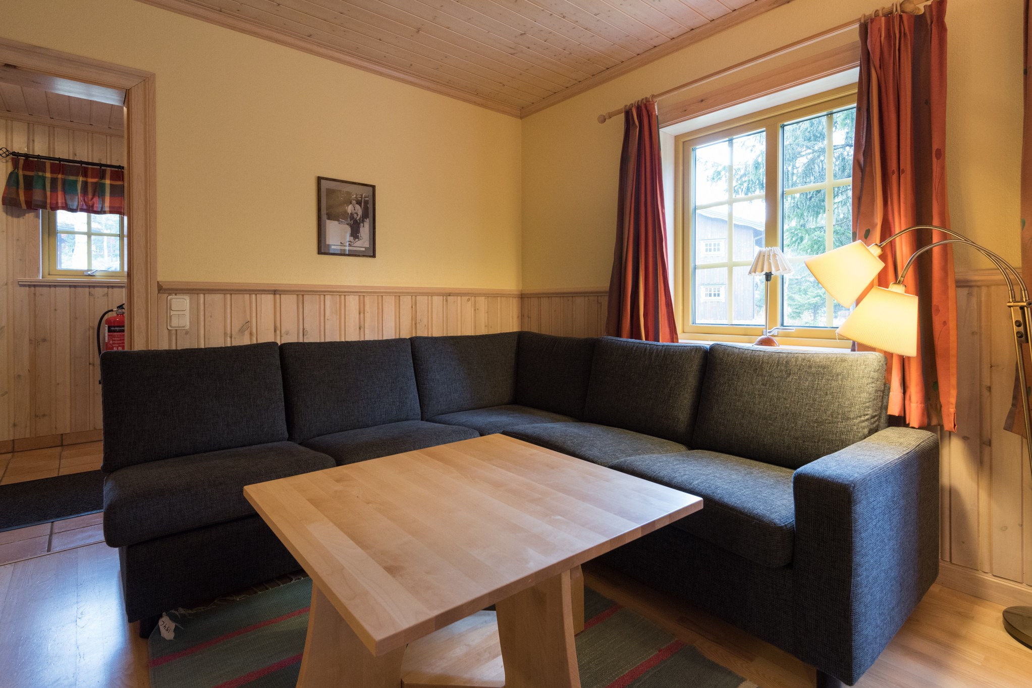 En hyggelig stue med en mørkegrå sektionssofa og et lyst sofabord i træ skaber den perfekte stemning til at planlægge dit sommerferie Østrig eventyr. Væggene med gule paneler og de orange gardiner tillader naturligt lys at oversvømme rummet, mens en gulvlampe giver ekstra belysning.