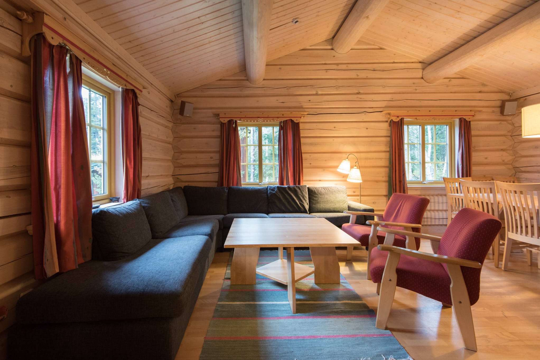 En hyggelig bjælkehyttestue i Saalbach byder dig velkommen med trævægge og loft. Den har en stor mørkegrå sektionssofa, to røde lænestole, et sofabord i træ og et spisebord med stole. Sollyset filtrerer ind gennem vinduerne med røde gardiner – perfekt efter en dag med skiferieeventyr.