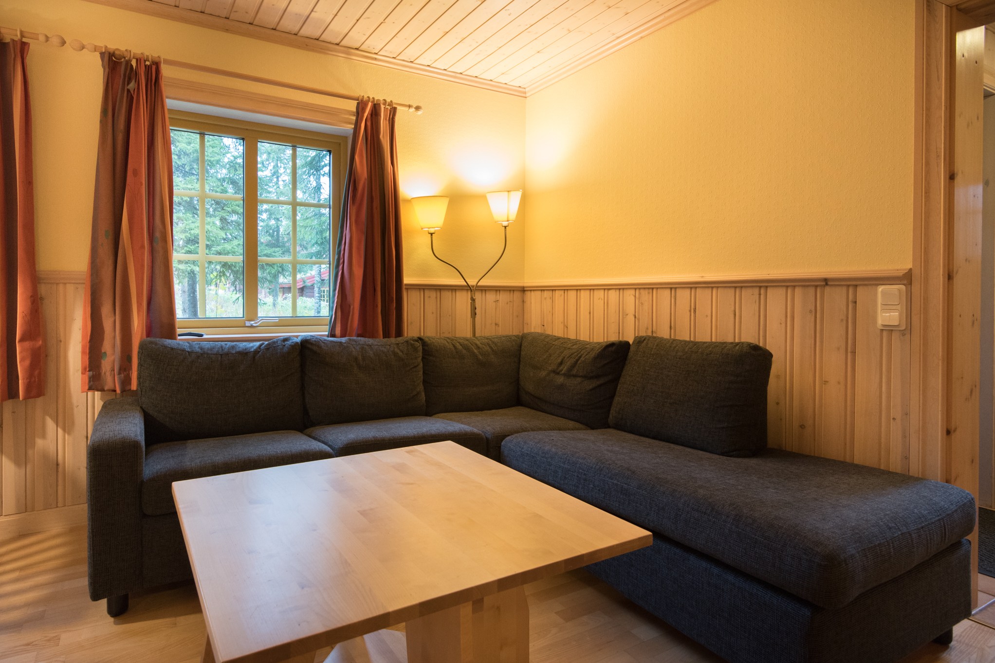 En hyggelig stue med en mørkegrå L-formet sofa, et sofabord i træ og en varm gul væg fremkalder charmen ved Westendorf. To vægmonterede lamper oplyser rummet, mens røde gardiner indrammer et vindue med udsigter, der minder om en skiferie i alpin skønhed.