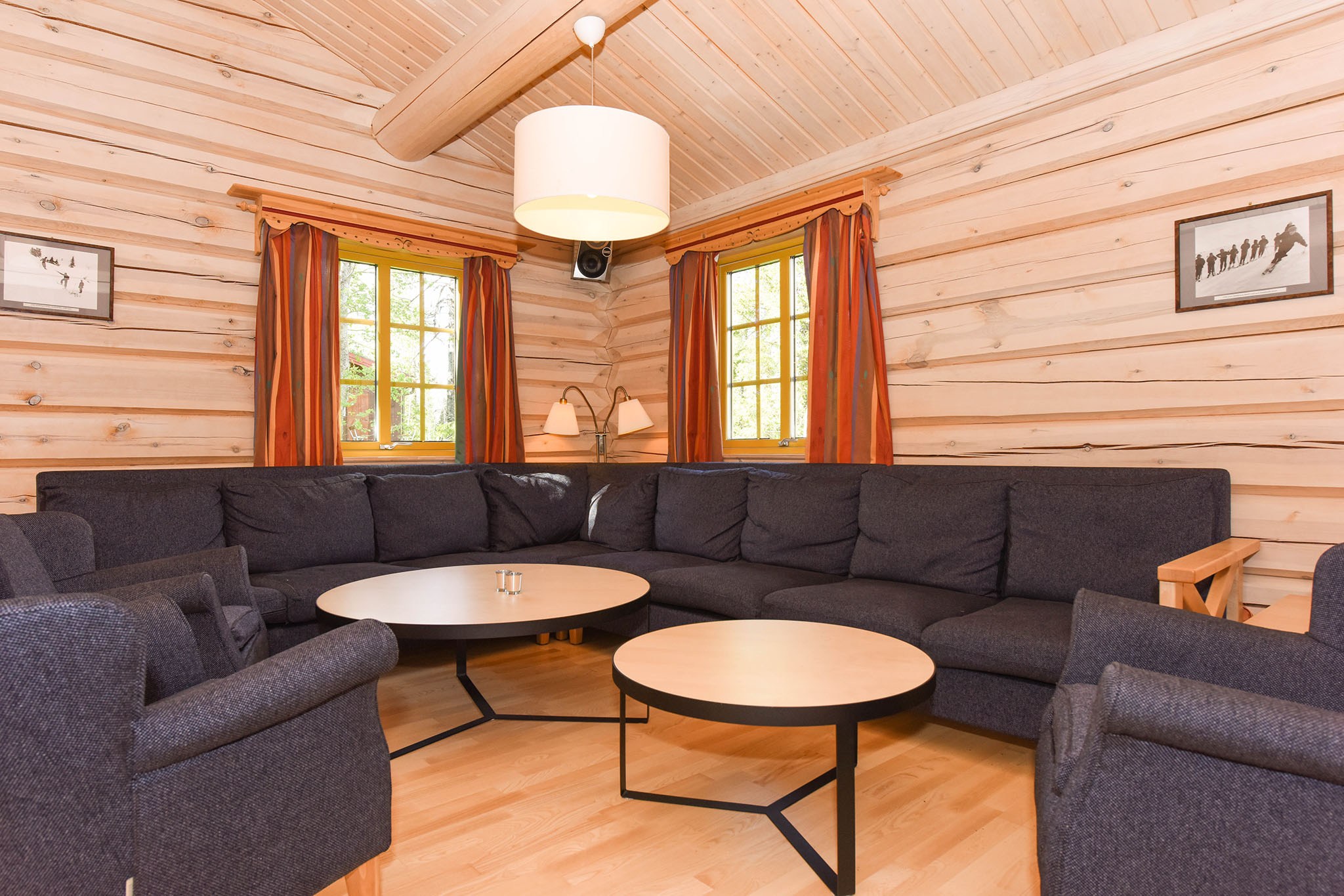 En hyggelig stue med trævægge og gulv, der minder om et skiferie-retræte, med en stor mørk sofa i L-form, to runde sofaborde og to lænestole. Store vinduer med røde gardiner giver naturligt lys. En loftlampe og indrammede billeder pryder rummet.