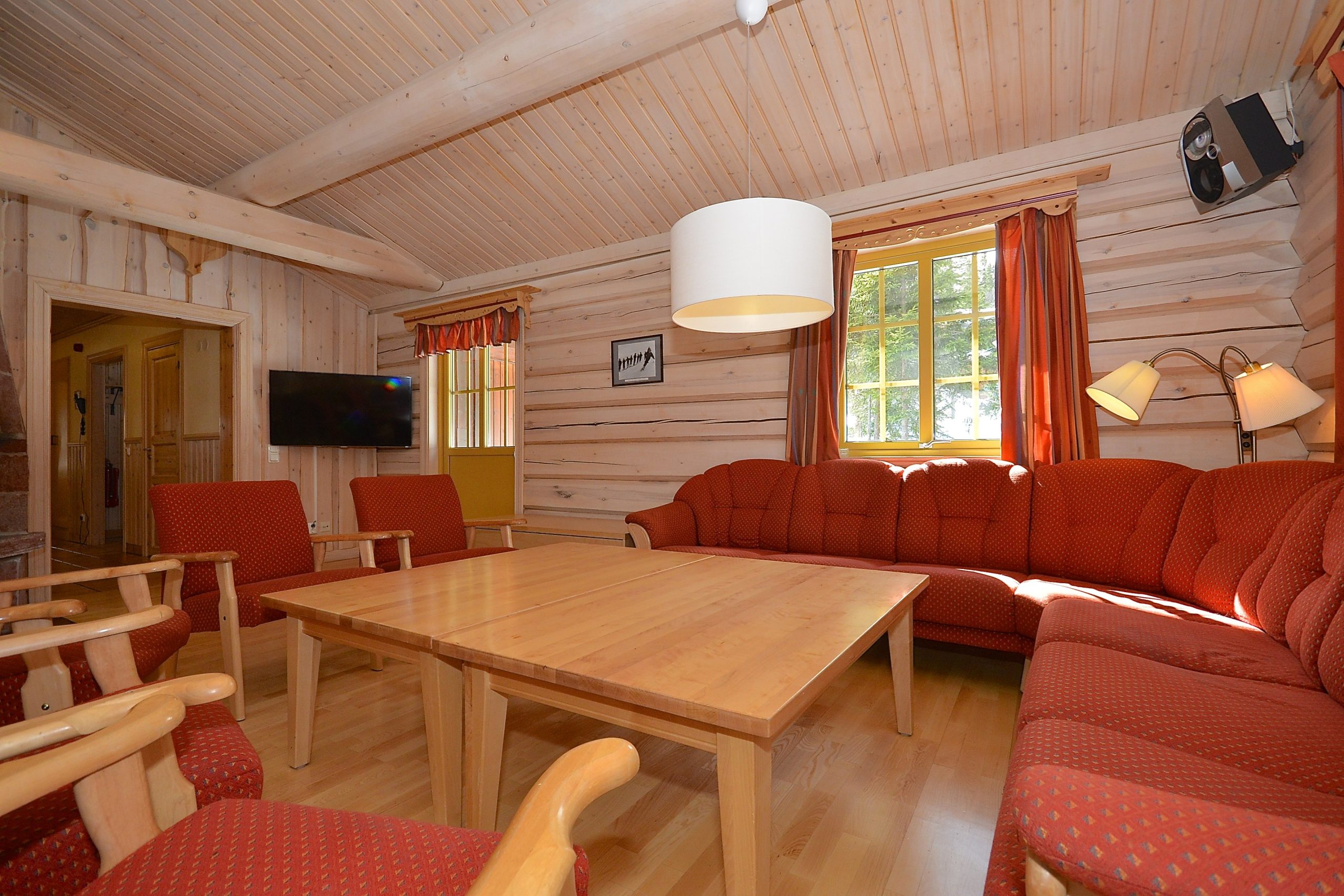 En hyggelig stue med træinteriør, der minder om en Saalbach-hytte, har røde polstrede sofaer og lænestole omkring et stort sofabord i træ. Et fladskærms-tv er monteret på væggen, og en pendel hænger fra loftet. Sollyset filtrerer gennem vinduet.