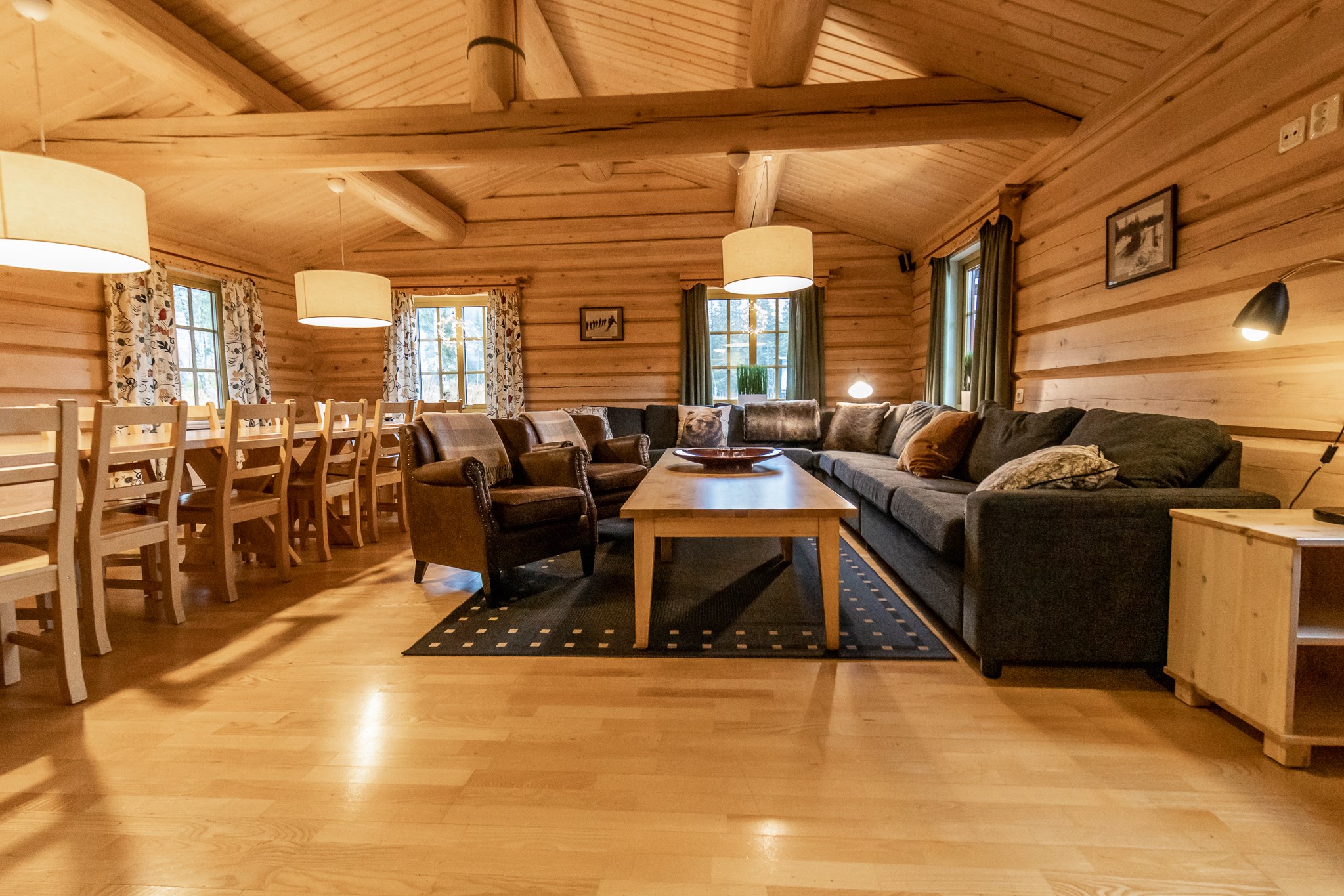 Et hyggeligt bjælkehytteinteriør med trævægge og loft, perfekt til en sommerferie Østrig-flugt. Der er et langt spisebord med stole og en stor L-formet sofa med lænestole omkring et sofabord i træ. Varm belysning og vinduer med blomstergardiner skaber en indbydende atmosfære.