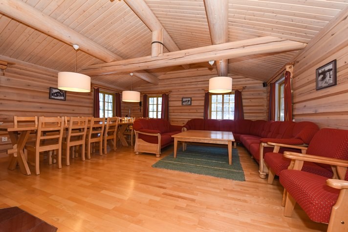 Interiøret i den hyggelige bjælkehytte, der minder om en charmerende Wagrain skiferie, har trævægge og loft. Et langt spisebord med træstole sidder til venstre, mens en rød sektionssofa og matchende stole skaber en indbydende krog til højre. Varm belysning fra hængende lamper lyser op i rummet.