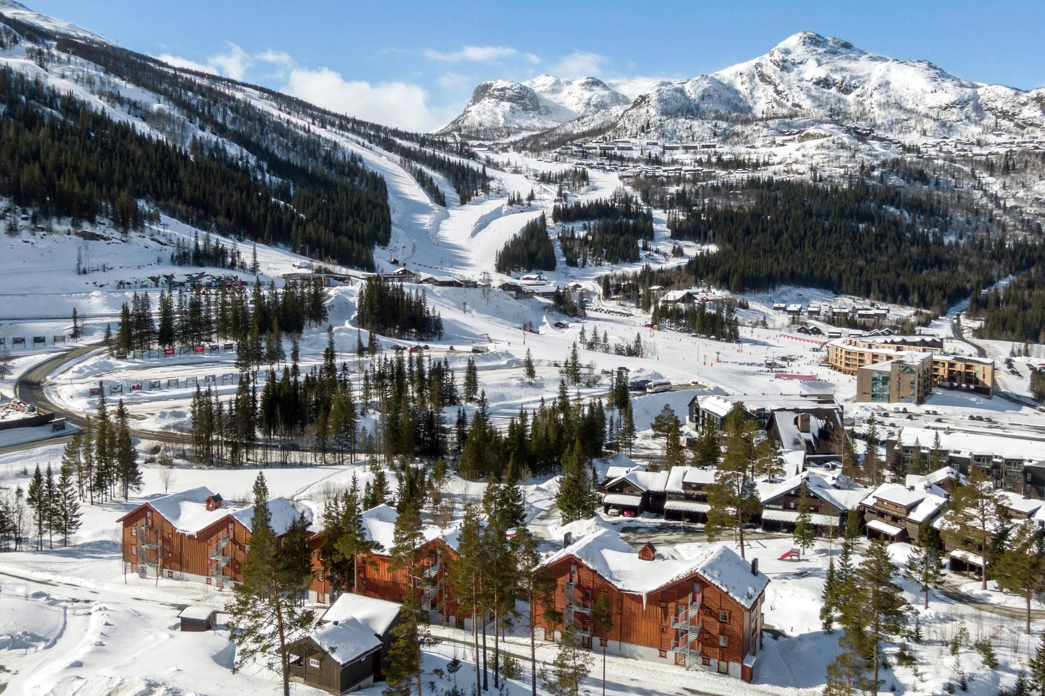 Et naturskønt vinterlandskab med et snedækket skisportssted i Saalbach med rustikke træhytter i forgrunden. Skiløjperne og stoleliften er sat mod skovklædte bakker og sneklædte bjerge under en klar blå himmel, og fanger den perfekte skiferiestemning.