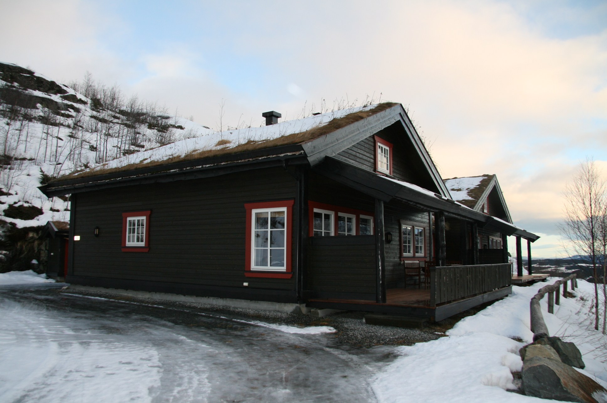 En træhytte med græsklædt tag er omgivet af sne, perfekt til en skiferie. Det har rødt trimmede vinduer og en veranda, beliggende ved siden af en stenet bakke. Himlen er delvist overskyet, hvilket skaber en fredfyldt vinterscene, der er ideel for skirejseentusiaster.